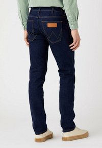 Vaqueros de mezclilla azul oscuro con corte slim, costuras naranjas visibles, dos bolsillos traseros y un parche de cuero marrón en la cinturilla.