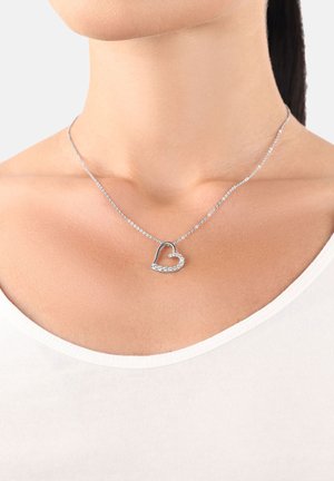 Collier en forme de cœur en argent avec de petits cristaux porté sur le cou d'une femme sur un haut blanc à encolure dégagée.