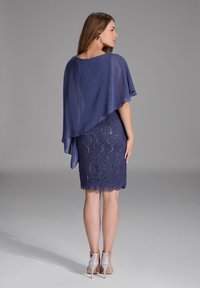 Robe marine avec un design en dentelle et un ourlet festonné, accompagnée d'un caplet léger et fluide. Présente des talons argentés et des détails subtils de brillance.