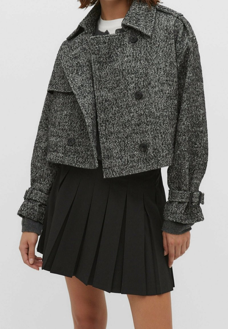 Femme portant une veste courte grise à chevrons avec des détails de boutons et une jupe plissée noire.