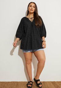 Svart, texturerad blus med V-ringning och knytdetalj, med puffärmar. Matchas med ljusa denimshorts och svarta sandaler.