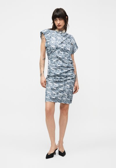 Γυναίκα που φοράει ένα μπλε φόρεμα με σχέδια μέχρι το γόνατο, με κοντά μανίκια τύπου καπελού και περιδέραιο από μαργαριτάρια, στέκεται μπροστά σε ένα απλό ανοιχτό φόντο.