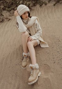 Botas de tobillo beige con borde de piel, suela resistente y ojales metálicos. Llevadas con un suéter crema y una bufanda, sentada en un suelo arenoso.