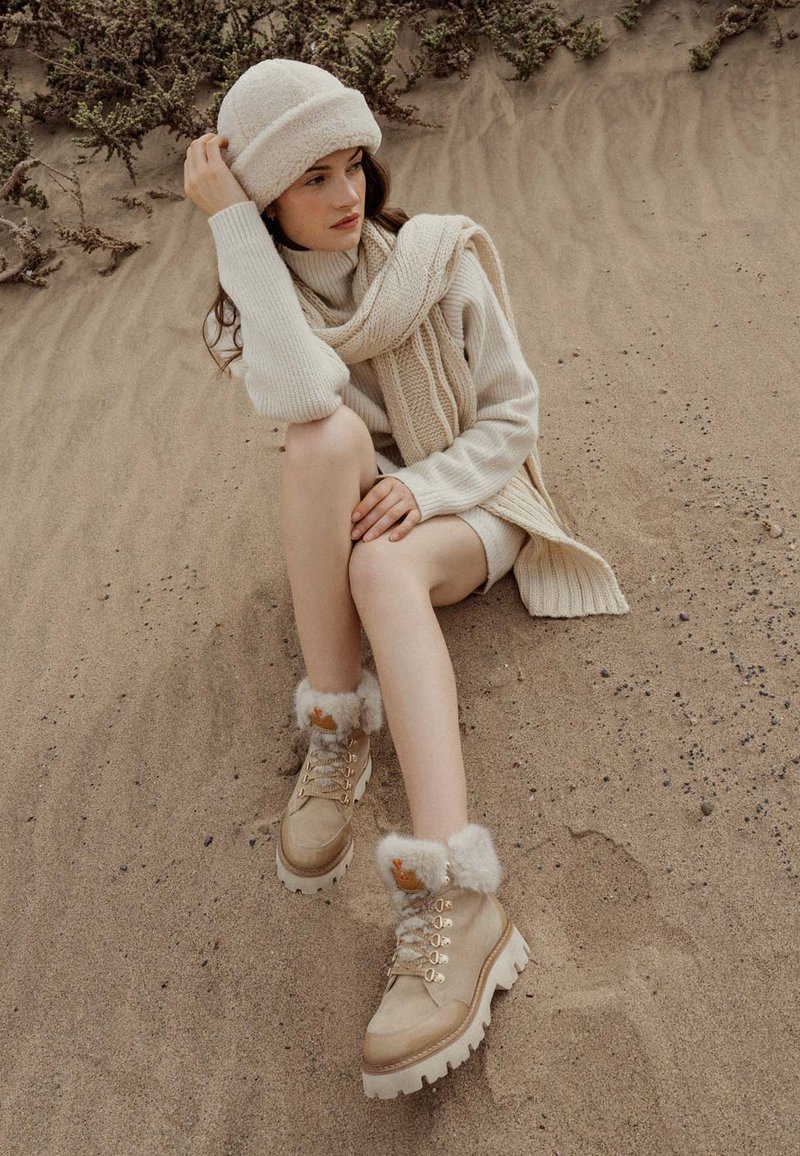 Botas de tobillo beige con borde de piel, suela resistente y ojales metálicos. Llevadas con un suéter crema y una bufanda, sentada en un suelo arenoso.
