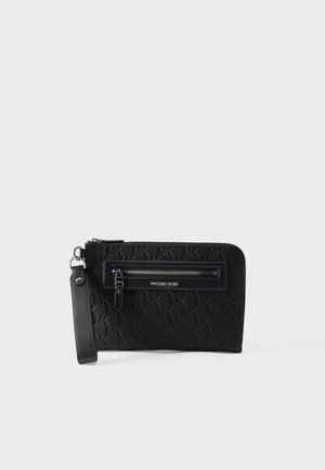 Pochette noire Michael Kors avec motif MK embossé, poche zippée à l'avant et bandoulière attachée sur fond clair.