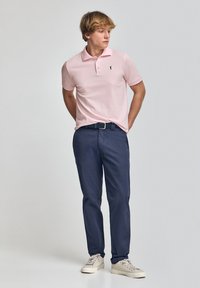 Rosa kortærmet polo skjorte med et sort logo, parret med navy bukser og beige sneakers, stående mod en neutral baggrund.