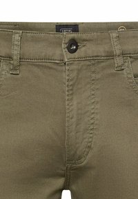 Vergrößerte Ansicht von olivgrünen Hosen mit Knopfverschluss, Gürtelschlaufen und sichtbarem Markenschild im Inneren des Bunds.