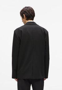Crni moderni sako s jednim prorezom straga, strukturiranim ramenima i glatkim materijalom. Minimalistički dizajn, bez vidljivih metalnih dijelova ili uzoraka.