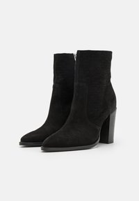 Paire de bottines en daim noir avec talons blocs et fermetures éclair latérales, présentant une couture subtilement texturée sur la partie supérieure.