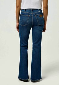 Jeans de denim com boca de sino em lavagem azul escura. Apresenta costura nos bolsos, metalurgia e um patch de couro texturizado próximo à cintura.