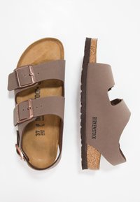Sandalias Birkenstock marrones con dos correas y hebillas, plantilla de corcho, suela de goma, lados abiertos y logo grabado en el lateral.
