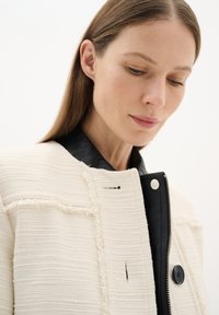 Giacca testurizzata color avorio con collare in pelle nera, dotata di zip frontale, chiusura con bottoni e bordi sfrangiati per un tocco di originalità.