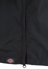 Fekete vízálló dzseki anyag sima textúrával. Elöl cipzárral ellátott, alján Dickies logós folttal. Egyszerű kialakítás.