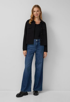Zwarte cropped jas met voorzakken, gedragen met hoog gewaagde, wijde blauwe jeans en stevige zwarte schoenen, vooraanzicht tegen een lichte achtergrond.