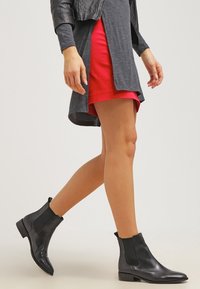Bottines en cuir noir avec un design épuré, des panneaux latéraux élastiques et un petit talon, portées avec un haut gris et une jupe rouge.