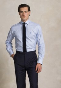 Gaiši zils šaha raksta krekls ar klasisku apkakli, savienots ar tumšām pielāgotām biksēm un struktūrētu melnu kaklasaiti. Redzama niansēta logotipa detaļa.