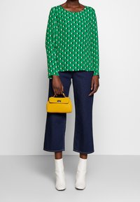 Grüne Bluse mit weißen Blattmustern, kombiniert mit dunkelblauen Jeans mit weitem Bein. Hält eine gelbe Handtasche. Weiße Stiefeletten vervollständigen den Look.