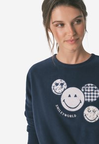 Marineblå sweatshirt med farverige cirkulære lapper med blomster- og ternede mønstre samt et centralt smilende ansigt design. Blød tekstur.