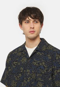 Camicia a maniche corte con un motivo astratto blu e verde, realizzata in tessuto leggero. Presenta un colletto a punta e due tasche sul petto.