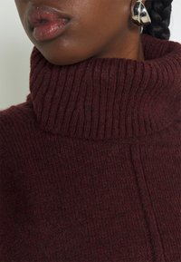 Maglione a collo alto in maglia bordeaux con trama a coste e dettaglio a punto contrastante, abbinato a un orecchino argentato lucido.