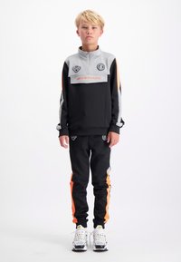 Zwarte en grijze trainingspak met oranje accenten. Het jack met rits heeft logo's; de broek heeft zijstrepen. Witte sportschoenen maken de look af.