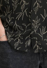 Chemise noire à boutons avec un motif de feuilles crème, fabriquée en tissu léger, avec une coupe décontractée et des détails de texture subtils.