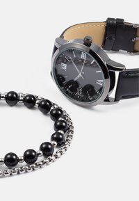 Czarny zegarek z rzymskimi cyframi, srebrnymi elementami i skórzanym paskiem, obok czarnej bransoletki z koralików z srebrnymi akcentami.