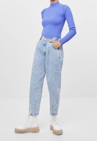 Jeans de tiro alto en azul claro con un ajuste holgado y tobillos ajustados, combinados con una camiseta de manga larga de canalé en morado y botas blancas voluminosas.