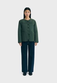 Giacca verde trapuntata con scollatura tonda, bottoni frontali e due tasche, abbinata a jeans scuri a gamba larga e scarpe nere.