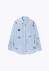 Chemise boutonnée bleu clair à rayures verticales blanches, à manches longues, avec une poche poitrine et des motifs colorés brodés représentant un soleil, une fleur et des formes géométriques.
