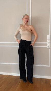 Lächelnde Frau steht drinnen und trägt ein beiges One-Shoulder-Oberteil mit Rüsche und schwarze, weit geschnittene Hose mit Nadelstreifen, Hände in den Taschen.