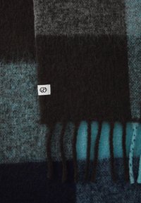 Gebreide wollen deken in tonen van teal, blauw, grijs en zwart, met gefronste randen en een klein wit label met een logo.