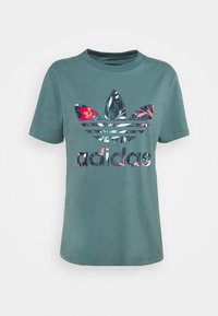 Turkos bomull t-shirt med korta ärmar, med en blommönstrad Adidas-logotyp och tre horisontella ränder under logotypen.
