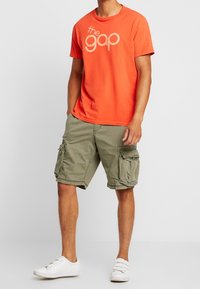 Oranssi puuvillainen t-paita, jossa on "the gap" -logo, yhdistetty oliivinvihreisiin cargo-shortseihin, joissa on useita taskuja, ja valkoisiin lenkkareihin.