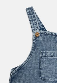 Salopette di jeans in un colore blu sbiadito, con spalline regolabili, una tasca frontale e dettagli in legno per la chiusura. Design in tessuto testurizzato.