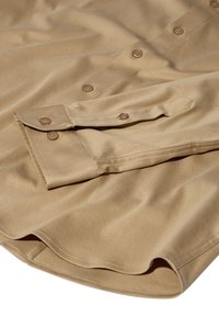 Beige langærmet skjorte med en glat tekstur, som har runde brune knapper og en manchet med knaplukning.