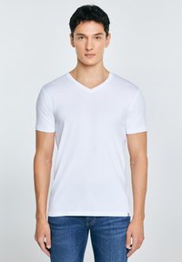 Joven con cabello corto y oscuro, vistiendo una camiseta blanca lisa de cuello en V y jeans azules, de pie frente a un fondo claro.