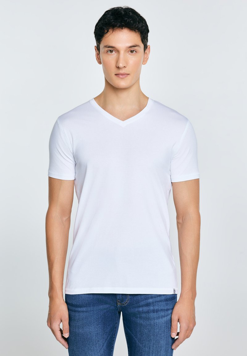 Joven con cabello corto y oscuro, vistiendo una camiseta blanca lisa de cuello en V y jeans azules, de pie frente a un fondo claro.