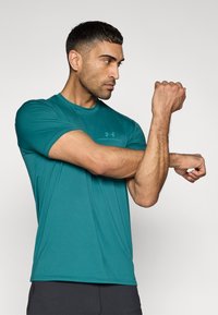 Τυρκουάζ αθλητική μπλούζα με κοντά μανίκια και λαιμόκοψη σε στρογγυλή γραμμή, κατασκευασμένη από λείο ύφασμα. Διαθέτει διακριτικό λογότυπο στο στήθος. Το μοντέλο κάμπτει το χέρι του.