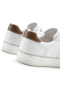 Weiße Leinwand-Sneaker mit braunem Akzent an der Ferse. Merkmale sind eine Gummisohle, gestickte Details und ein klassisches, flaches Design.