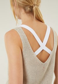 Débardeur en tricot beige avec un dos en V et des bretelles blanches croisées. Présente un tissage texturé et un petit détail brodé.