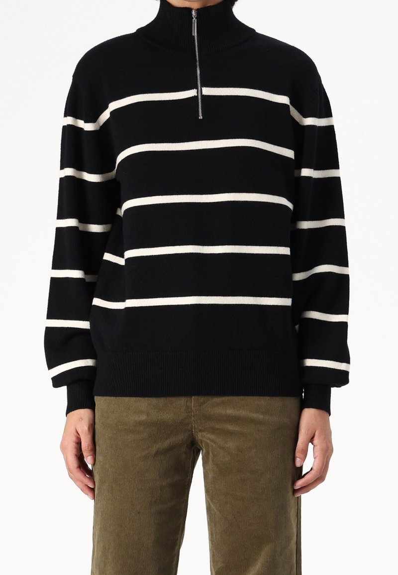 Pull à rayures noires et blanches avec col montant et fermeture à moitié zippée. Tissu tricoté avec poignets et ourlet côtelés. Porté avec un pantalon en velours côtelé olive.
