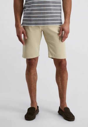 Mann trägt beige Shorts, dunkelbraune Slipper und ein grau-weiß gestreiftes Hemd, steht vor einem schlichten hellen Hintergrund.