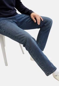 Jeans di denim in un lavaggio blu scuro con taglio dritto. Tessuto di cotone con una sottile trama, dettagli cuciti, abbinati a sneakers bianche.