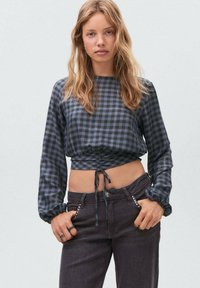 Plava karo bluzica skraćena s dugim rukavima, nabranim prednjim dijelom i detaljem svezanja, u kombinaciji s tamnoplavim trapericama koje imaju ukrašene džepove.