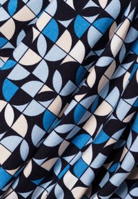 Bawełniana tkanina z geometrycznym nadrukiem, w którym na przemian występują niebieskie, jasnoniebieskie, kremowe i granatowe kształty w wzorze siatki. Teksturowana powierzchnia.