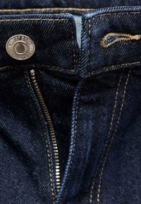 Los jeans de mezclilla azul oscuro presentan un botón plateado y una cremallera, con costuras y bolsillos de hilo dorado en contraste.