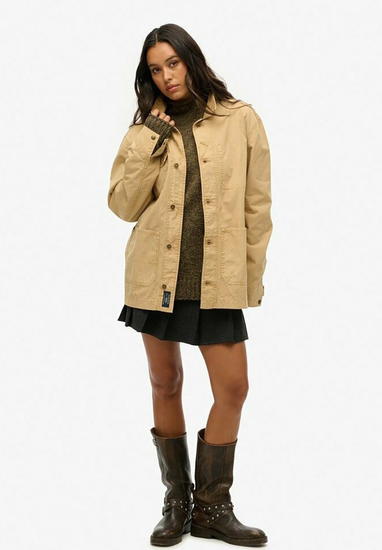 Chaqueta de algodón beige con dos bolsillos frontales, cierre de botón y un corte relajado, combinada con un suéter oscuro y una falda negra. Botas marrones hasta la rodilla.