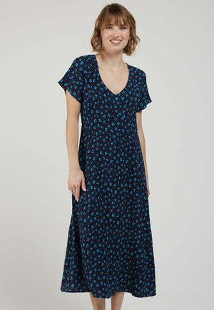Femme souriante, portant une robe midi bleu marine à manches courtes avec un motif floral bleu, debout devant un fond clair uni.