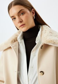 Beige ullkappa med en krage av konstgjord päls, med en dubbelsidig design och en ljus knapp, bärs över en mörk ribbad polotröja.